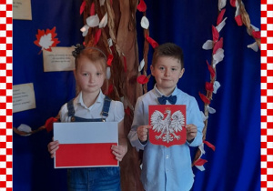 Marcelinka i Maks w odświętnym ubraniu trzymają w rączkach godło i flagę Polski, w tle dekoracja okolicznościowa